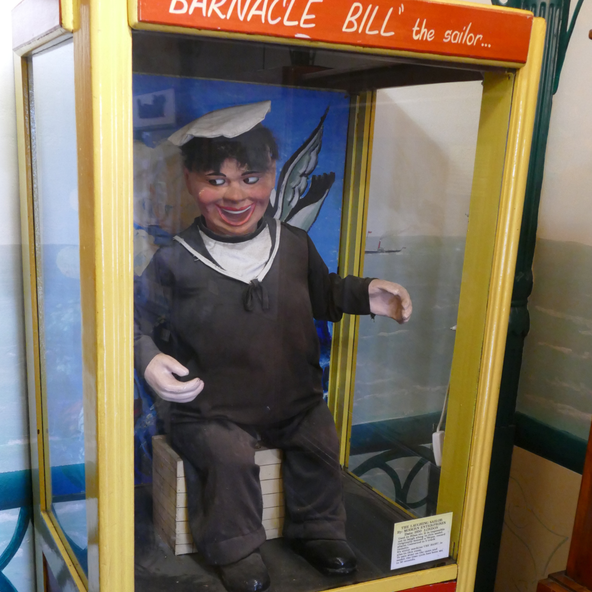 Vintage penny arcade display at of Old Pier Penny Arcade showing a sailor automaton seated inside a yellow coin‑operated cabinet.